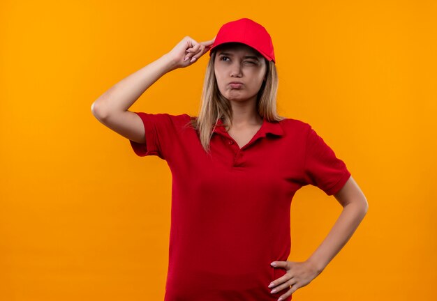 Kijkend naar kant verward jong leveringsmeisje die rood uniform en pet dragen die vinger aan hoofd zetten die op oranje achtergrond wordt geïsoleerd