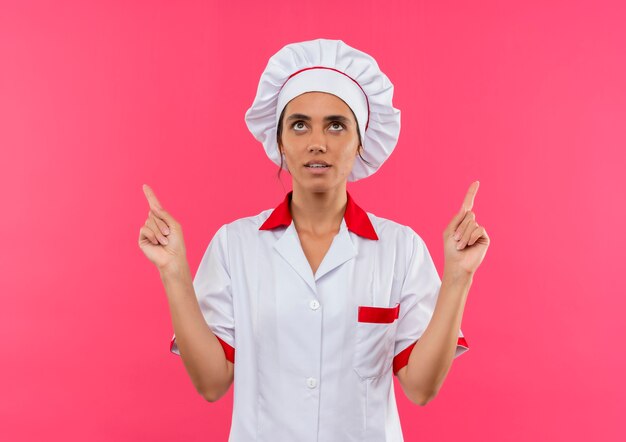 Kijkend naar omhoog jonge vrouwelijke kok dragen chef-kok uniform wijst vinger omhoog op geïsoleerde roze muur met kopie ruimte