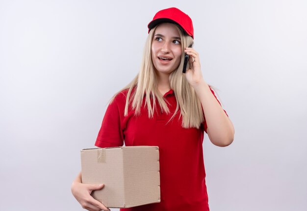 Kijkend naar zijbezorging jonge vrouw, gekleed in rood t-shirt en pet in tandheelkundige beugel met doos en pseakes op telefoon op geïsoleerde witte muur