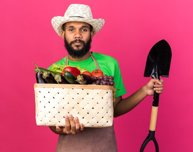 Kijkende camera jonge tuinman afro-amerikaanse man met tuinhoed met groentemand met spade