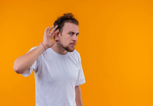 Kijkende jonge kerel die wit t-shirt draagt dat luistergebaar op geïsoleerde oranje achtergrond toont