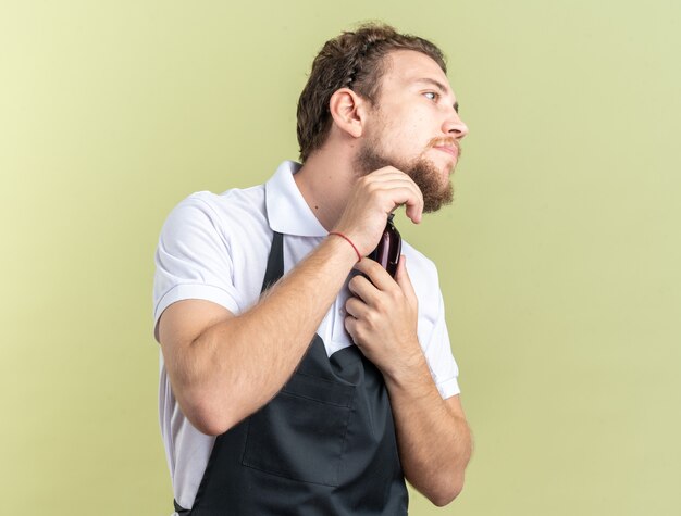 Kijkende jonge mannelijke kapper die een uniforme trimbaard draagt met een tondeuse geïsoleerd op een olijfgroene muur