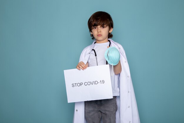 Kind jongen houden stop covid hashtag in witte medische pak en grijze jeans op blauwe muur