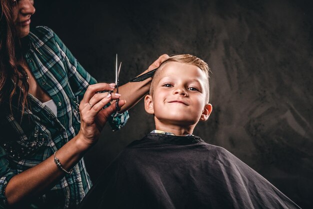 Kinderkapper met een schaar snijdt kleine jongen tegen een donkere achtergrond. Tevreden schattige kleuterjongen die kapsel krijgt.