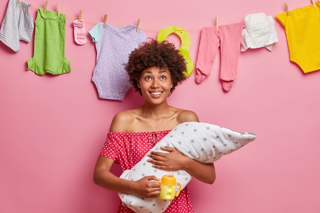 Kinderopvang, gelukkig ouderschap, gezin en liefde concept. Positieve lachende vrouw vormt met slaperige pasgeborene na het voeden, melkfles houdt, geniet van het moederschap, staat binnen. Kleine baby in moedershanden