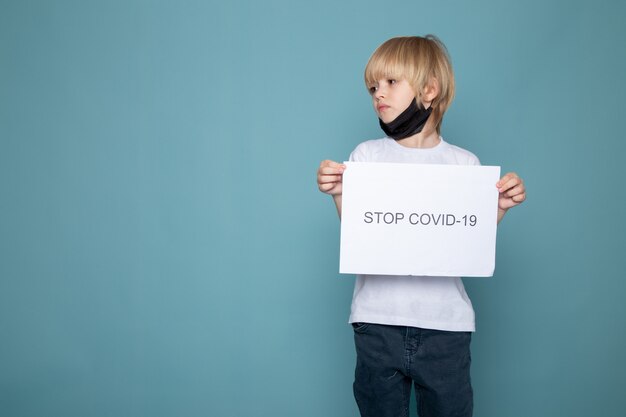 Kindjongen met blond haar in wit t-shirt en spijkerbroek samen met stop covid hashtag op blauwe muur