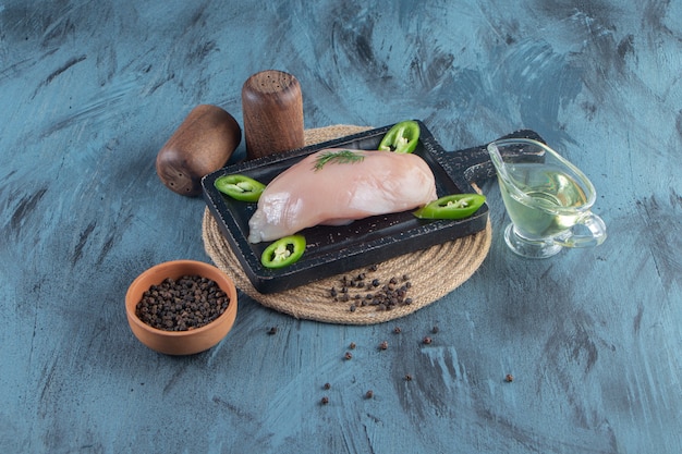 Kipfilet en gesneden peper op een bord op een onderzetter naast kruiden- en oliekommen, op het blauwe oppervlak. Premium Foto