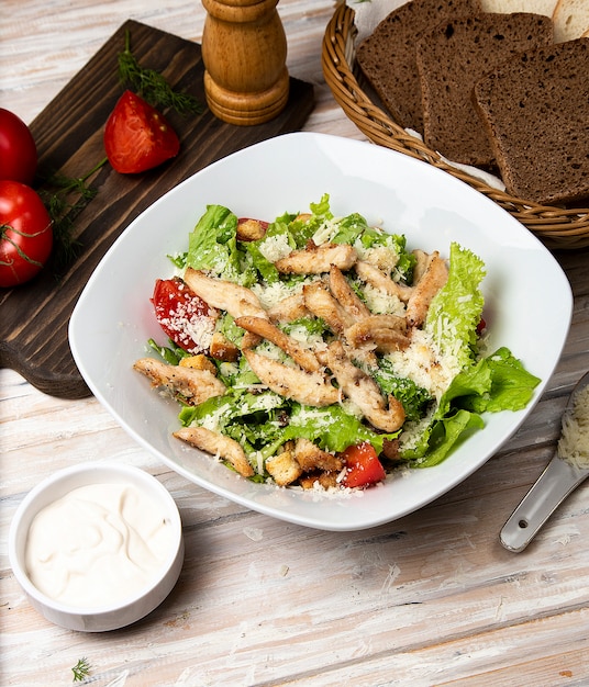 Kippen parmezaanse kaas caesar salade met sla, cherrytomaatjes in een witte kom, geserveerd met saus en brood.