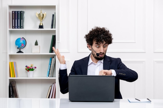 Klantenservice knappe krullende man in kantoorpak met computer en headset die naar klokken kijkt