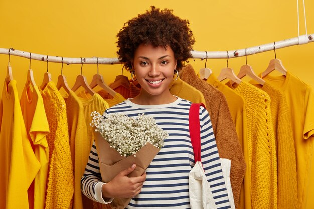 Kleding verkoop concept. Vrolijke gekrulde Afro-Amerikaanse vrouw in goed humeur, houdt mooi boeket, glimlacht breed, draagt boodschappentas, staat tegen kledingrekken