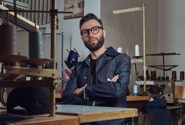 Kleermaker gekruiste armen zittend aan een tafel met naaimachine, kijkend naar een camera in een naaiatelier.