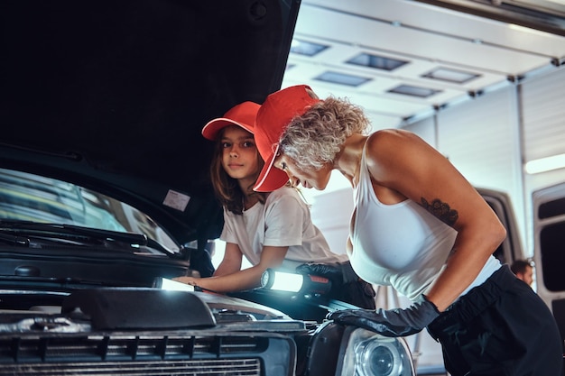 Klein lachend meisje is nieuwsgierig naar de motor van de auto bij de autoservice met haar moeder, die het werk van een man doet.