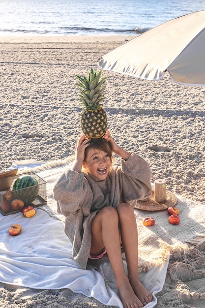 Gratis foto klein meisje aan de zandige kust bij een picknick met fruit