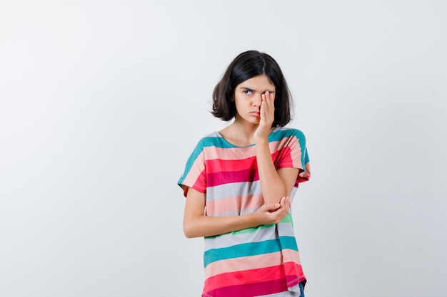 Klein meisje dat het oog bedekt met de hand terwijl ze de hand op de elleboog houdt, wegkijkt in een t-shirt en er slaperig uitziet, vooraanzicht.