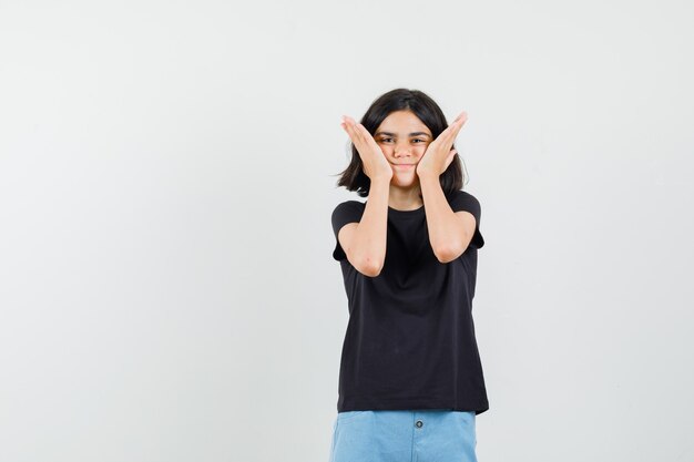 Klein meisje hand in hand op wangen in zwart t-shirt, korte broek en op zoek schattig, vooraanzicht.