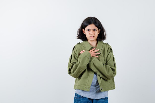 Klein meisje houdt gekruiste handen op de borst in jas, t-shirt, spijkerbroek en kijkt weemoedig, vooraanzicht.