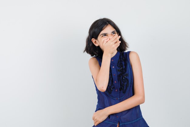 Klein meisje in blauwe blouse met hand op de mond terwijl lachen, vooraanzicht.
