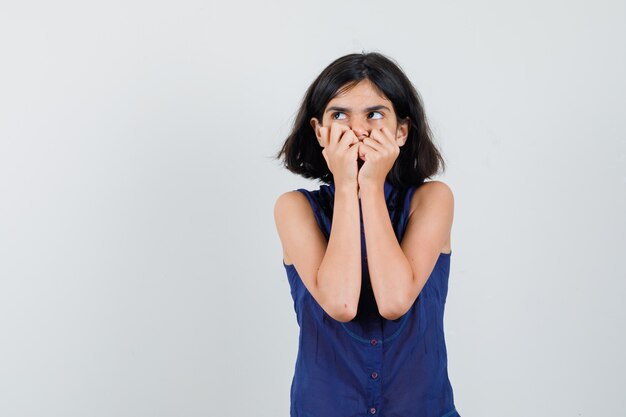 Klein meisje in blauwe blouse met vuisten op de mond en bang, vooraanzicht op zoek.