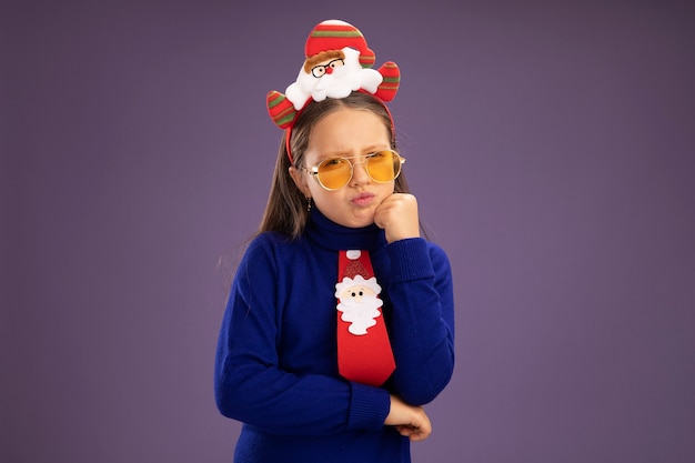 Klein meisje in blauwe coltrui met rode stropdas en grappige kerstrand op hoofd camera kijken met sceptische uitdrukking staande over paarse achtergrond