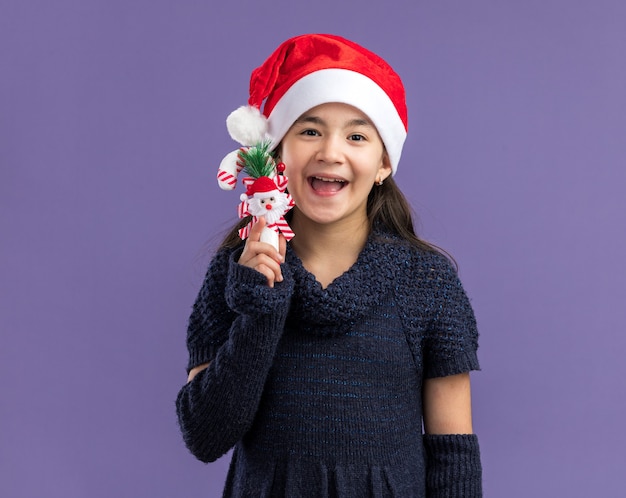 Klein meisje in een gebreide jurk met een kerstmuts met kerstsnoepgoed blij en vrolijk over de paarse muur