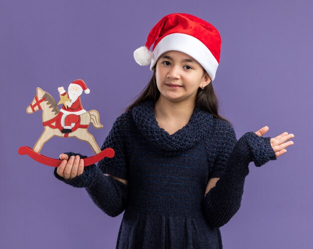 Klein meisje in een gebreide jurk met een kerstmuts met kerstspeelgoed met een glimlach op het gezicht over een paarse muur