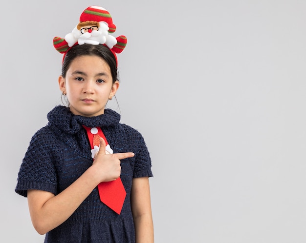 Klein meisje in gebreide jurk, gekleed in een rode stropdas met grappige kerstrand op het hoofd en kijkt met een ernstig gezicht dat met een uit-vinger naar de zijkant wijst