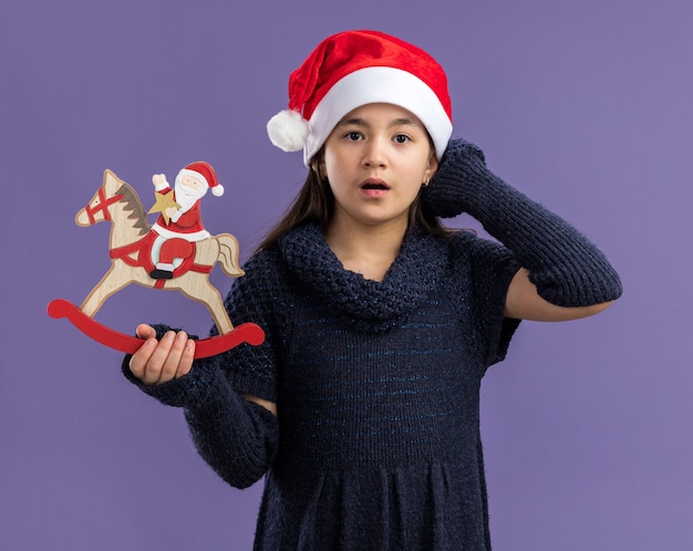 Klein meisje in gebreide jurk met een kerstmuts met kerstspeelgoed verbaasd over de paarse muur