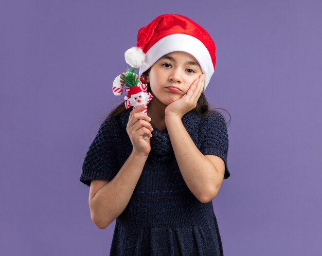 Klein meisje in gebreide jurk met kerstmuts bedrijf kerst candy cane op zoek verward met droevige uitdrukking