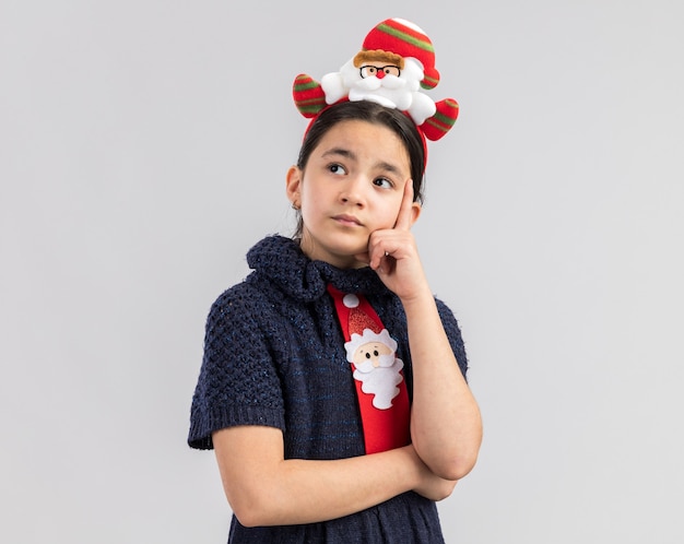 Klein meisje in gebreide jurk met rode stropdas met grappige kerst rand op hoofd kijken met peinzende uitdrukking denken
