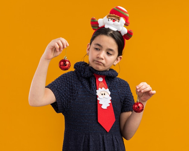 Klein meisje in gebreide jurk met rode stropdas met grappige rand op het hoofd met kerstballen die verward kijken en proberen een keuze te maken die over een oranje muur staat