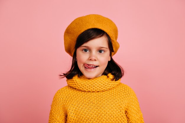 Klein meisje in gebreide trui staande op roze muur. kind poseren met tong uit.