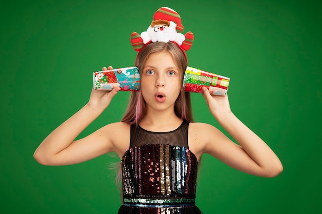 Klein meisje in glitter feestjurk en kerstman hoofdband met kleurrijke papieren bekers over haar oren en kijkt verbaasd over groene achtergrond