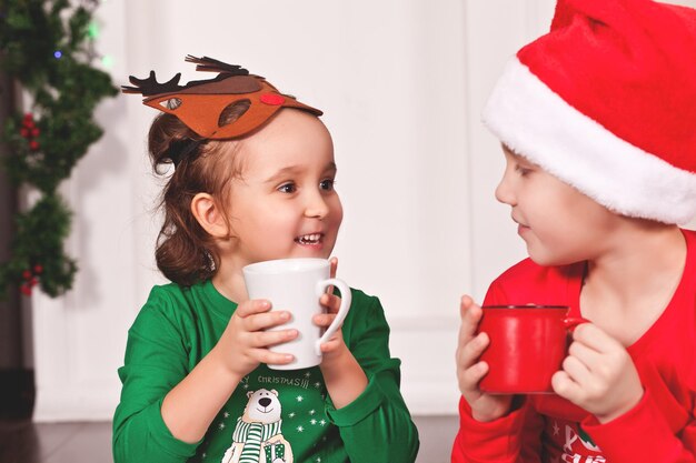 Klein meisje in grappig hertenmasker en jongenskind in kerstmuts en kerstpyjama die cacao drinken