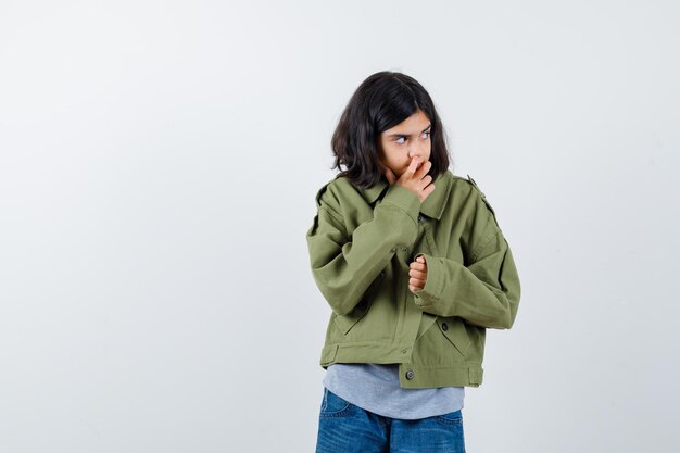 Klein meisje in jas, t-shirt, spijkerbroek die op haar nagels bijt terwijl ze wegkijkt en peinzend kijkt, vooraanzicht.