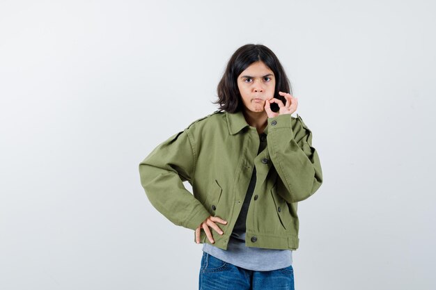 Klein meisje in jas, t-shirt, spijkerbroek met ok gebaar en zelfverzekerd, vooraanzicht.