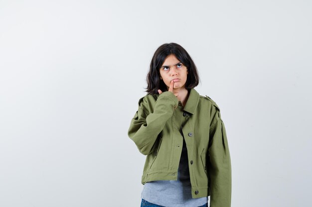 Klein meisje in jas, t-shirt, spijkerbroek staat in denkende pose en ziet er attent uit, vooraanzicht.