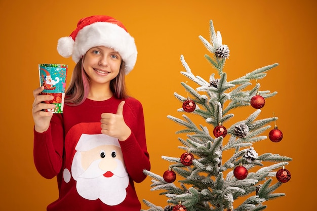 Klein meisje in kerstsweater en kerstmuts met kleurrijke papieren beker glimlachend duimen opdagen naast een kerstboom over oranje achtergrond