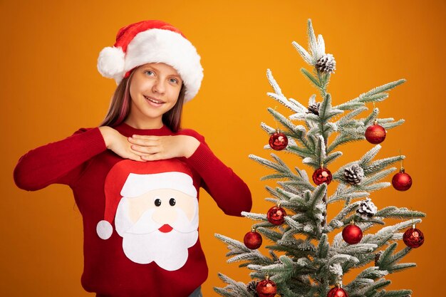 Klein meisje in kersttrui en kerstmuts blij en verrast glimlachend vrolijk hand in hand op haar borst zich dankbaar voelend naast een kerstboom over oranje achtergrond