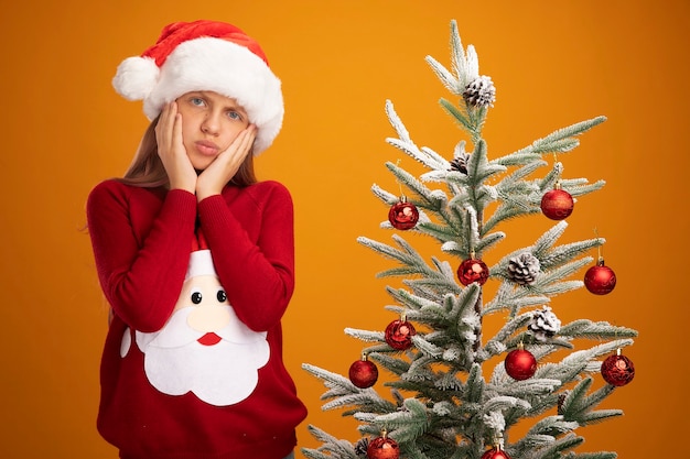 Klein meisje in kersttrui en kerstmuts kijkend naar camera verward en bezorgd naast een kerstboom over oranje achtergrond