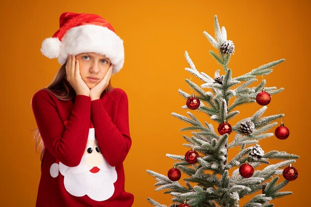 Klein meisje in kersttrui en kerstmuts kijkend naar camera verward en ontevreden naast een kerstboom over oranje achtergrond