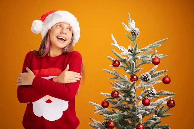 Klein meisje in kersttrui en kerstmuts knuffelt zichzelf gelukkig en vrolijk glimlachend naast een kerstboom over oranje achtergrond