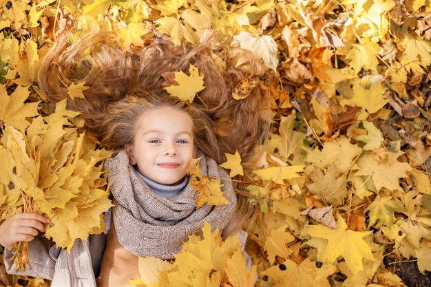 Klein meisje in mode kleding in herfst bos. Meisje liggend in een gele bladeren. Meisje dat bruine sjaal draagt en in camera kijkt.