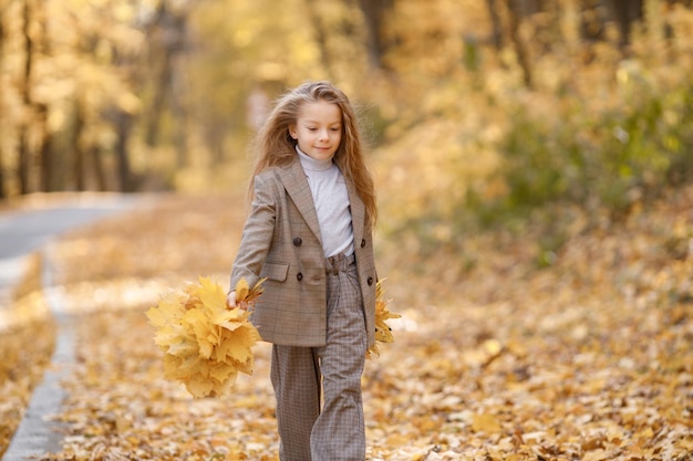 Klein meisje in mode kleding wandelen in herfst park. Meisje met een gele bladeren. Meisje dat bruin kostuum met jasje draagt.