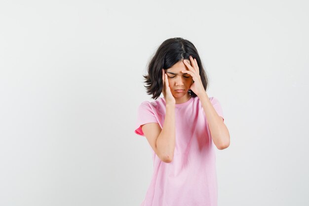 Klein meisje in roze t-shirt met sterke hoofdpijn en ziet er moe, vooraanzicht uit.