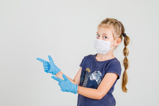 Klein meisje in t-shirt, handschoenen en masker wijst met twee vingers camera kijken.