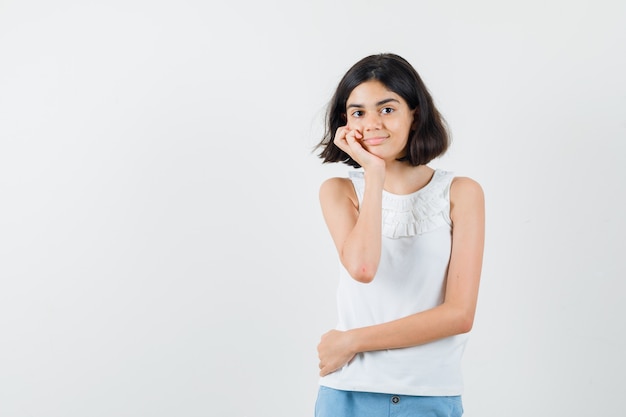 Klein meisje in witte blouse, korte broek staande in denken pose en op zoek verstandig, vooraanzicht.