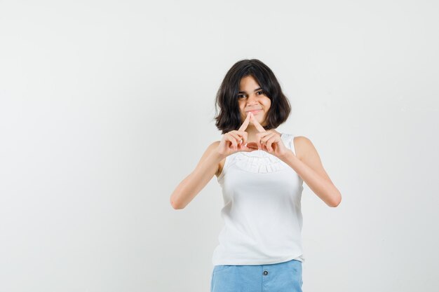 Klein meisje in witte blouse, korte broek vorm met vingers en op zoek vrolijk, vooraanzicht.