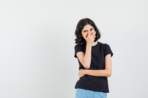 Klein meisje in zwart t-shirt, korte broek hand op de mond houden en op zoek gelukkig, vooraanzicht.