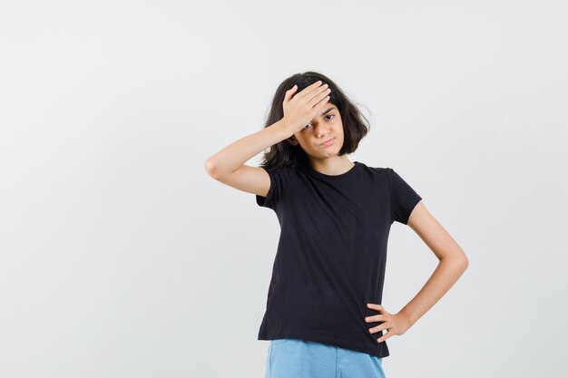 Klein meisje in zwart t-shirt, korte broek hand op het voorhoofd houden en spijtig, vooraanzicht op zoek.