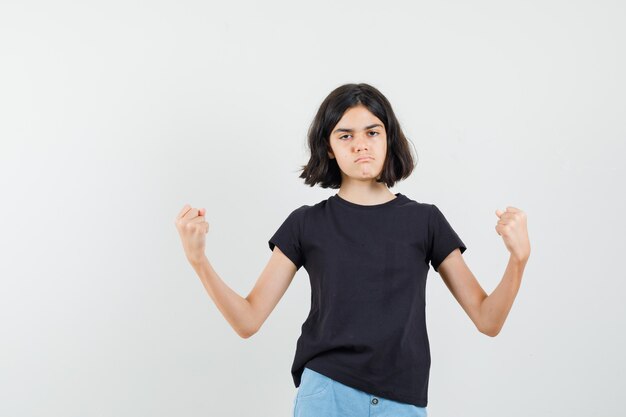 Klein meisje in zwart t-shirt, korte broek met succesgebaar en op zoek naar zelfverzekerd, vooraanzicht.